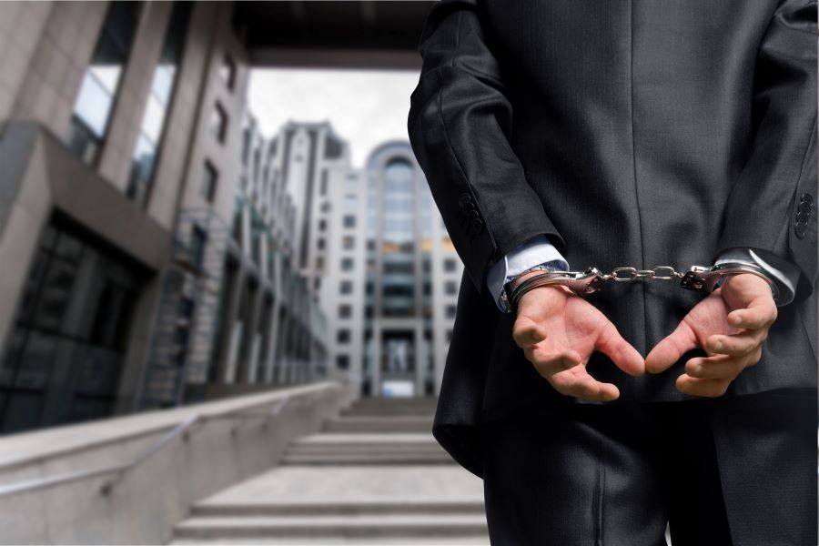 A man in a suit, his hands behind him in handcuffs, standing in front of a courthouse