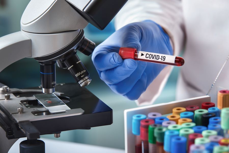 A scientist holds a vial labeled "COVID-19" in front of a microscope