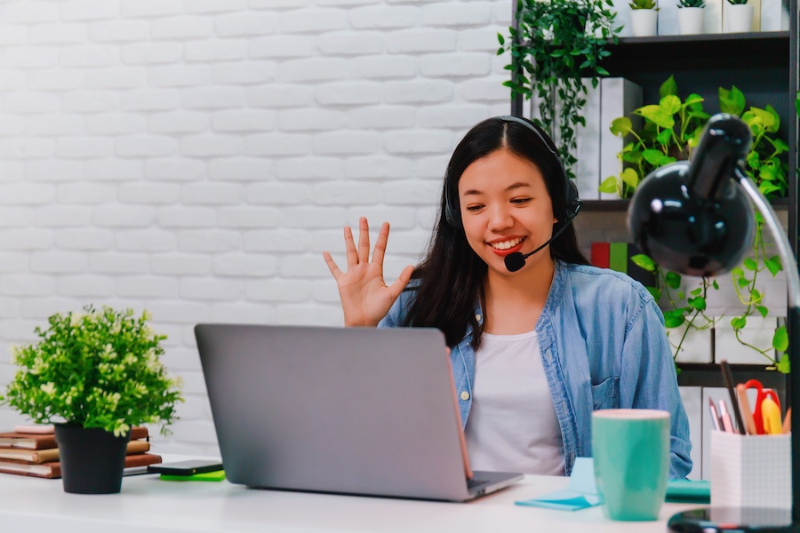 A home in a home office wears a headset microphone and waves to her laptop