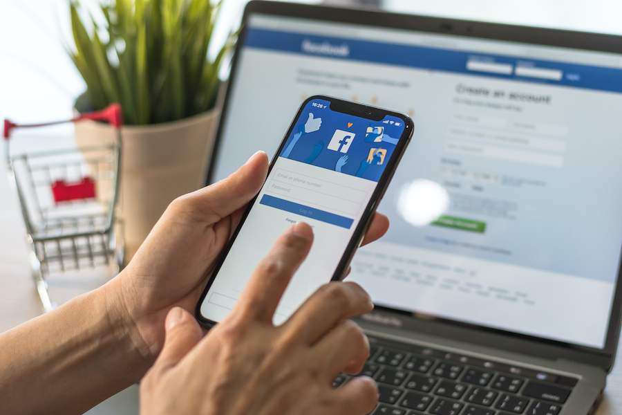 Hands hold a smartphone showing the log in screen for facebook while a laptop in the background also shows the login screen for facebook