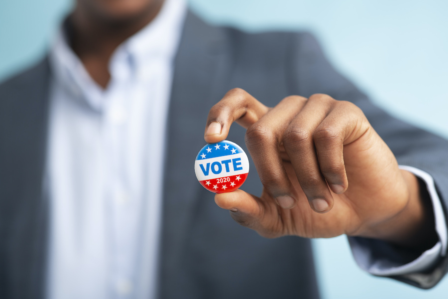 A man in a casual suit hold up a button that reads, "VOTE 2020"