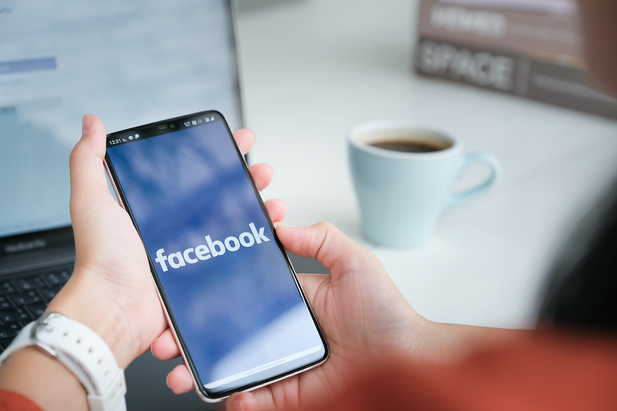 A person opens Facebook on a smartphone, a laptop and cup of coffee are visible in the background