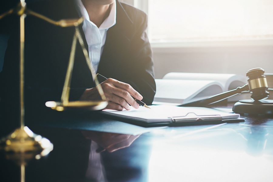 A person makes notes in a legal proceeding, a gavel and the scales of justice are visible on the desk