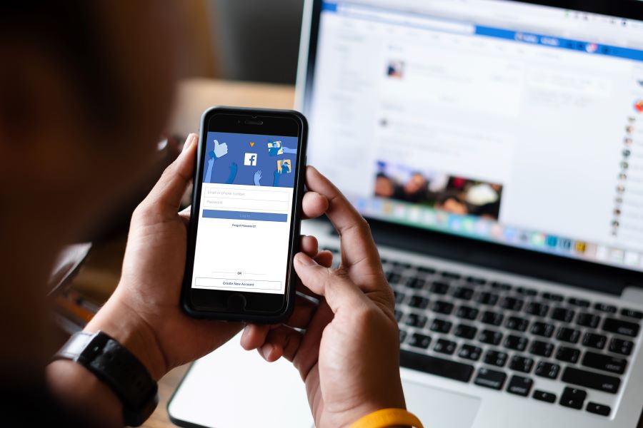 A person holds a smartphone in front of a laptop. Both are displaying Facebook's homepage.