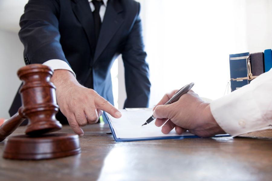 A hand signs a contract while a man in a suit points at the contract, there is a gavel on the table