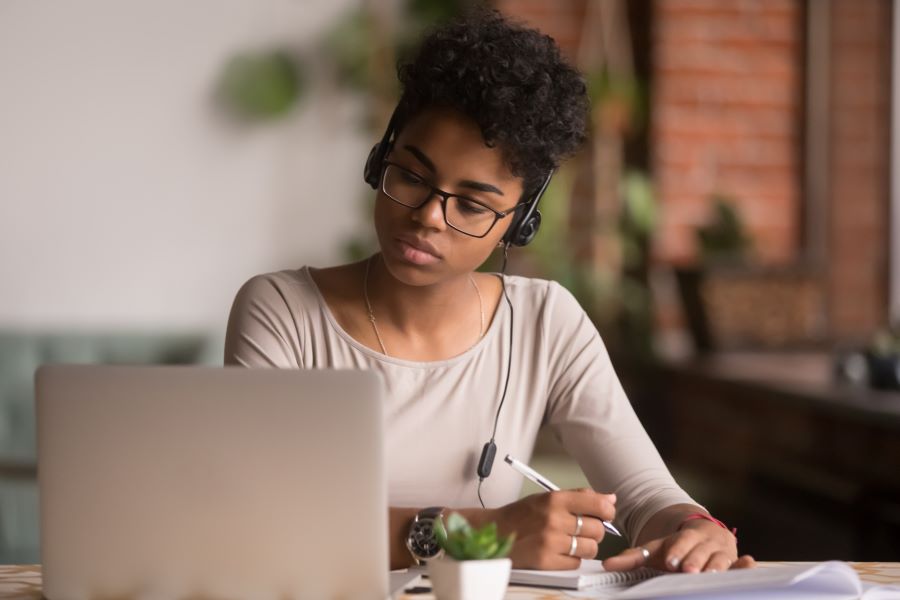 A woman wears headphones and takes notes while looking at a laptop