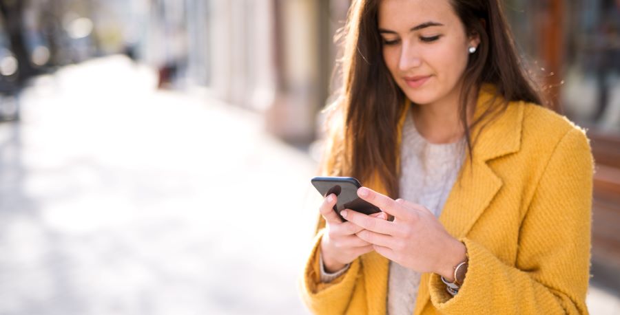 A woman in a yellow blazer is using a smartphone while standing on a city sidewalk