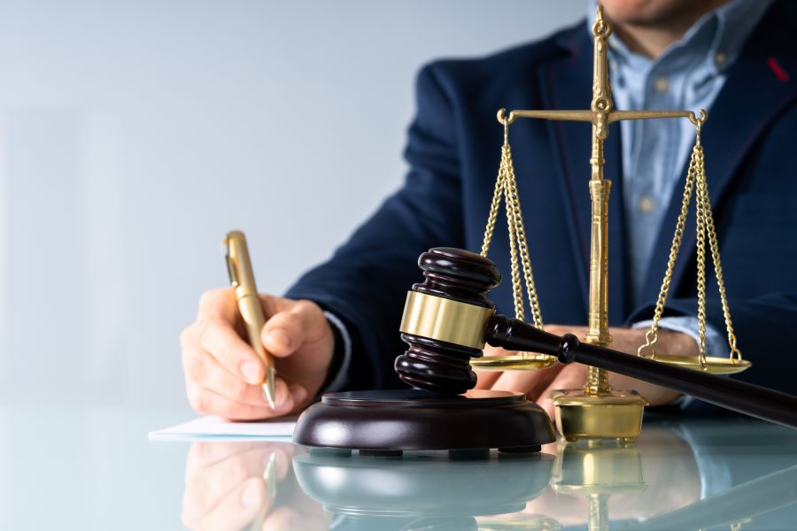 A man in a blue suit signs legal documents with a gavel and scales of justice in the foreground