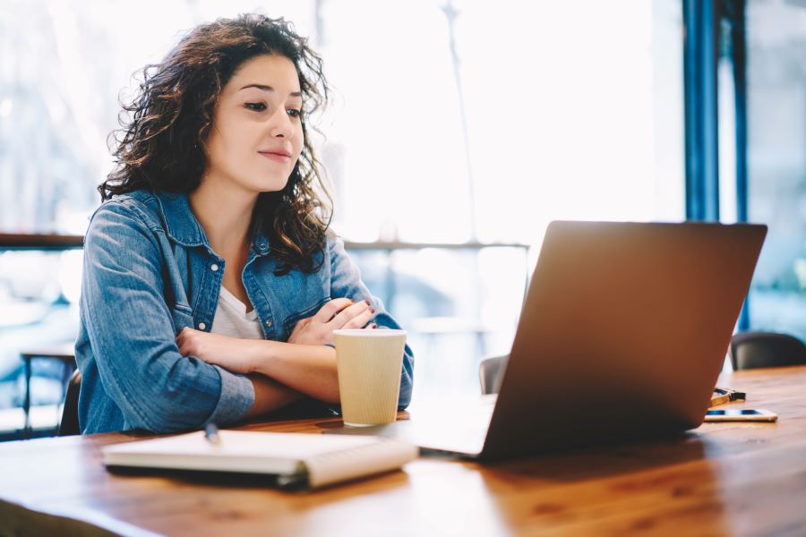 A woman in a jean jacket watches something on a laptop while seated in a coffee shop