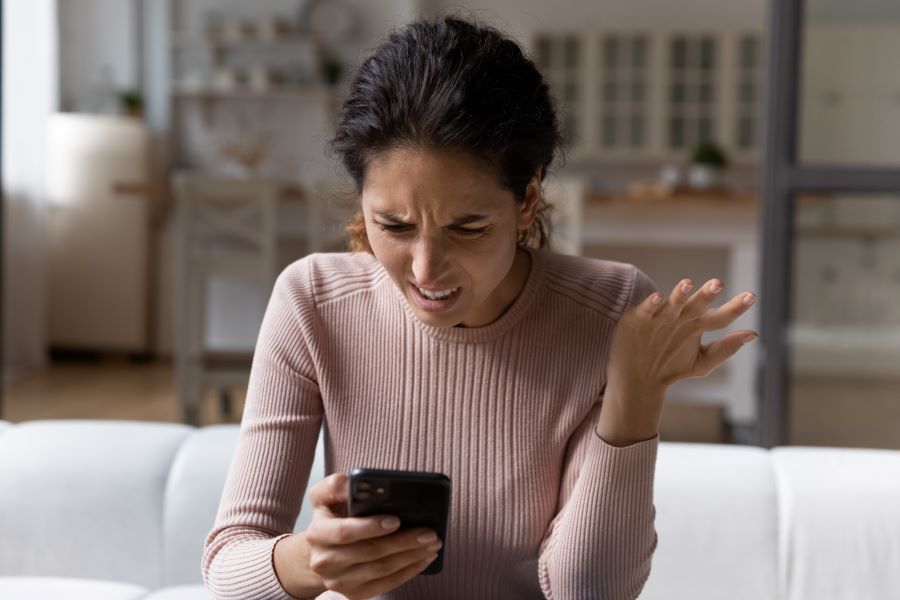 A woman seated on a couch looks at her phone, makes a disgusted face, and incredulous hand gesture