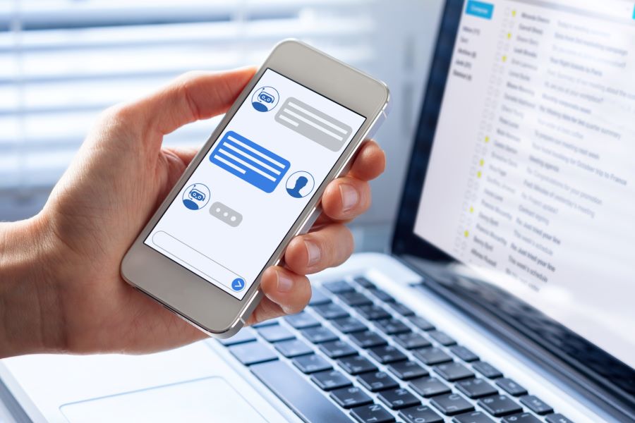 A hand holds a smartphone showing a chatbot conversation, in the background a laptop is open showing an email inbox