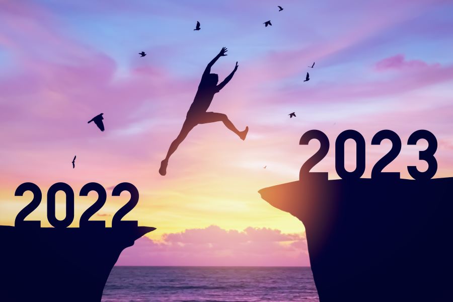 A silhouette of a man jumping from a cliff that is labeled 2022 to one that is labeled 2023, seascape at sunset in the background