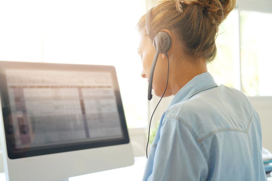 A woman in a blue shirt wears a headset telephone and looks at a computer screen