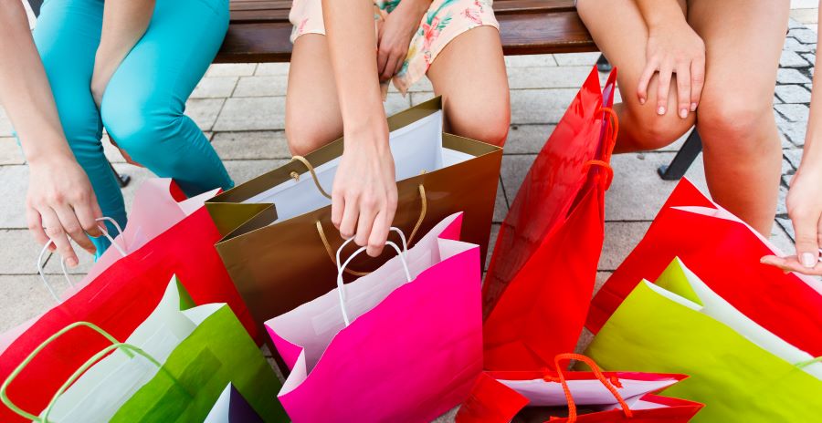 The kneews and hands of three people are visible as they sit on a bench and reach for 8 multicolored shopping bags